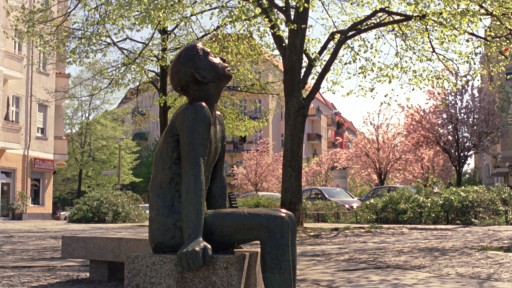 Auf einer Bank unter Bäumen reckt eine Metallstatue ihr Gesicht nach oben zur Sonne