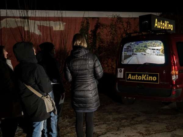 Menschen schauen auf die Heckscheibe eines roten Hauses, auf dem ein Film gezeigt wird. Auf dem Auto steht "AutoKino".