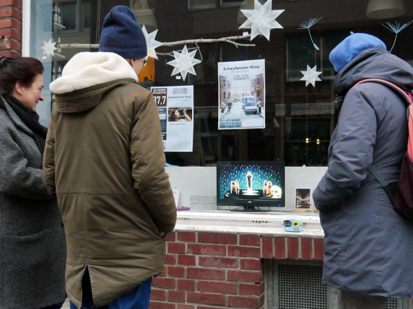 Drei Menschen stehen vor einem Ladenfenster. Auf einem Fernseher hinter der Glasscheibe wird ein Film gezeigt. Davor steht ein sehr kleines Radio.