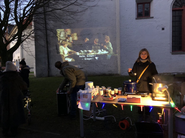 Vor einer Kirche steht ein Tisch, auf dem unter anderem Tassen und T&ouml;pfe stehen. Eine Frau hat eine Sch&ouml;pfkelle in der Hand. An der Kirchenwand ist ein Film projiziert.